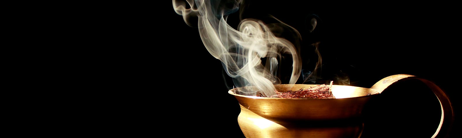 Photo of a golden goblet with handle, on a light brown surface, against a black background. The contents of the goblet are smoking, with trails of smoke going up and to the left of the image.