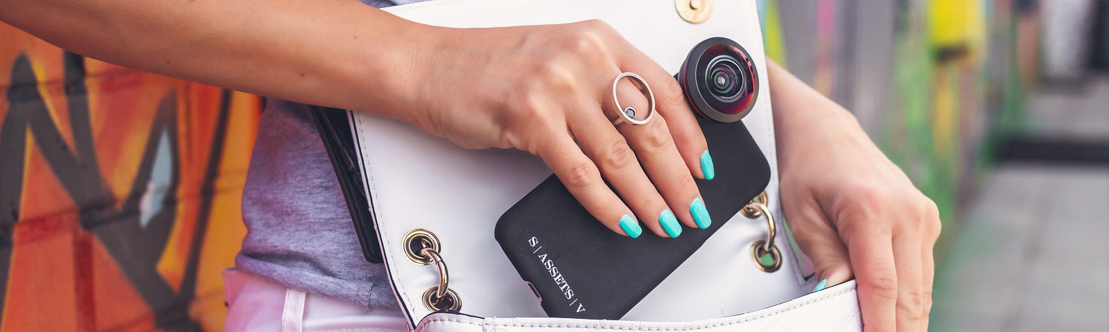 A white handbag is slung across a woman’s body. It is open and the woman is slipping her phone into it. She has turquoise nail polish on and behind her is a wall with red and green graffiti.