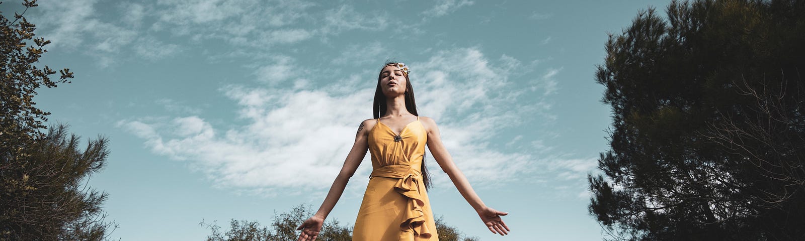 A holy woman in long flowing dress stands amid trees on a hill, spreading her arms wide, looking skyward