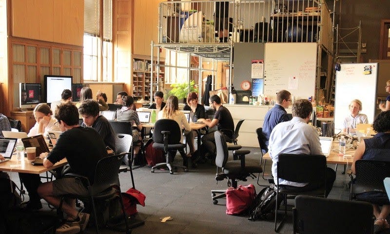 A photograph of about 20 people in conversation at different tables. Many are working on laptops.