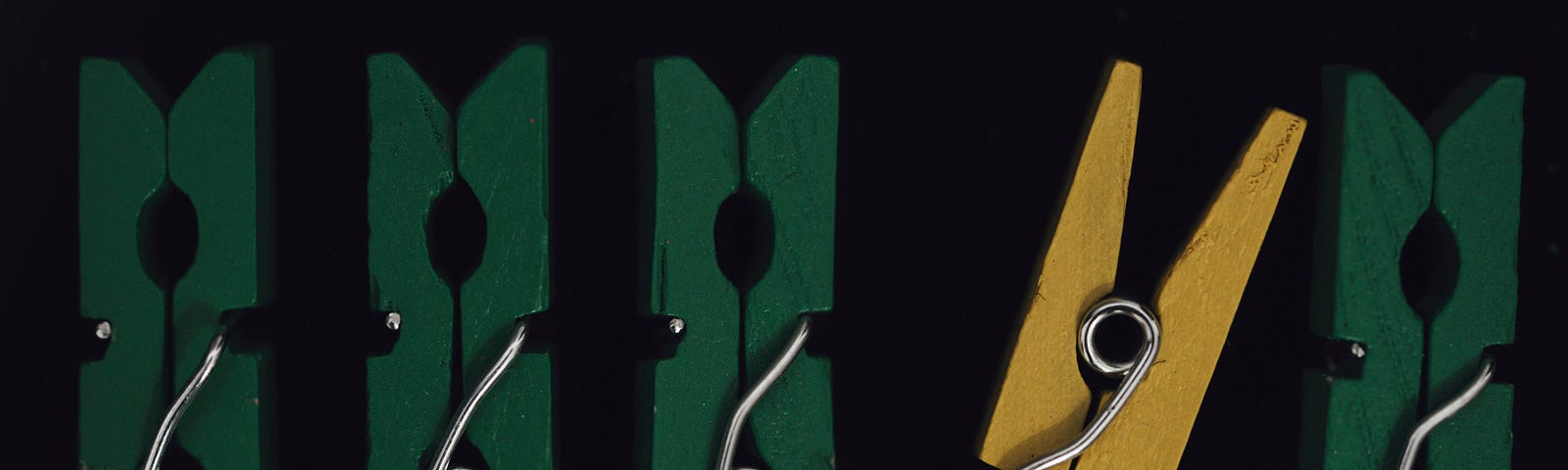 The image shows five clothespins against a black background. Four of the clothespins are green, while one is yellow. The yellow clothespin stands out and leans slightly against one of the green ones, highlighting the idea of being different within a group.