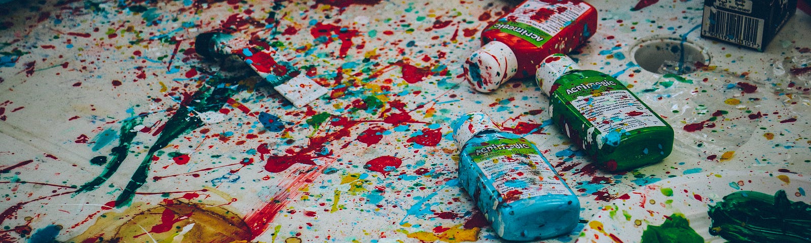 Image shows a cloth covered in paint splatters and smudges in yellow, light blue, red and yellow, with different bottles of paint lying on top of the cloth (also covered in splatter).