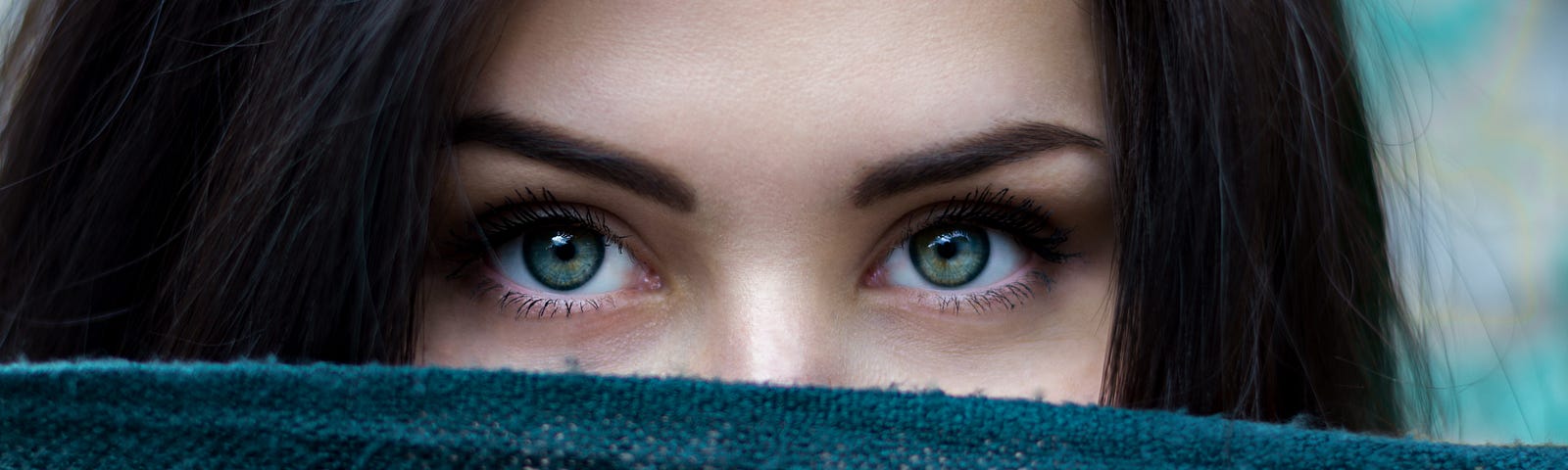 Dark-haired woman with big green eyes looking over a blue cloth that covers the bottom portion of her face.