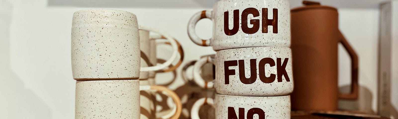 A side table, like a night stand, with five ceramic coffee mugs in two stacks (a stack of two and a stack of three). The coffee mugs in the-three stack each have a word emblazoned on the front in dark brown. The first words says “Ugh.” The second word says “Fuck.” The third word says “No.”