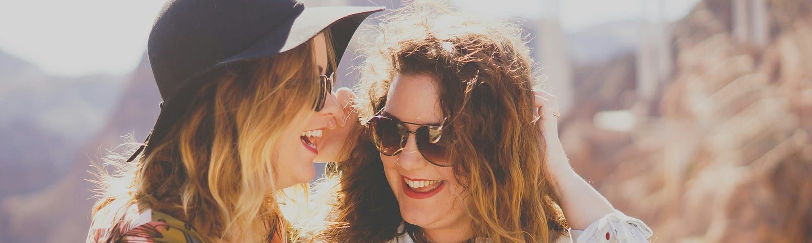 Two women one wearing a hat and the other leaning in and smiling to hear a secret.