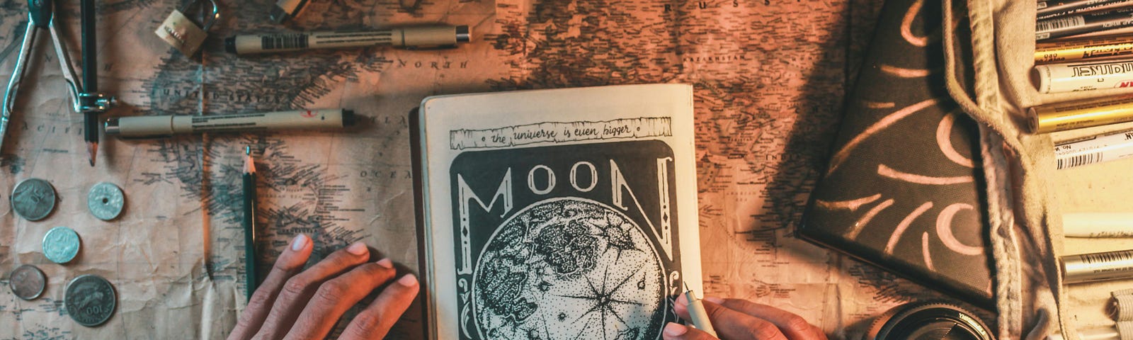 A notebook with a sketch of the moon on a desk surrounded by pens, maps, and other writing materials