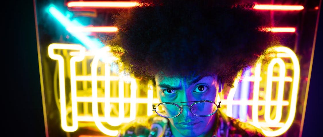 Man with afro and glasses wearing a funky shirt in a neon lit room looking at the camera with a curious expression