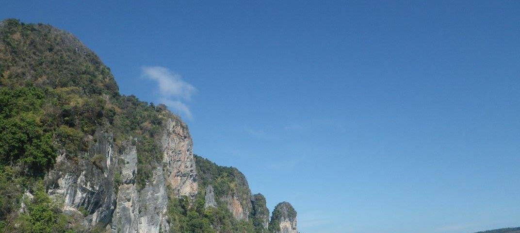 A photo of an island off the coast of Phuket Thailand in January 2016. The water is gradient from light to dark, left to right, a teal to icy blue. The large rocky island is casting no shadows as the day is bright and sunny. There is a small boat off to the left.