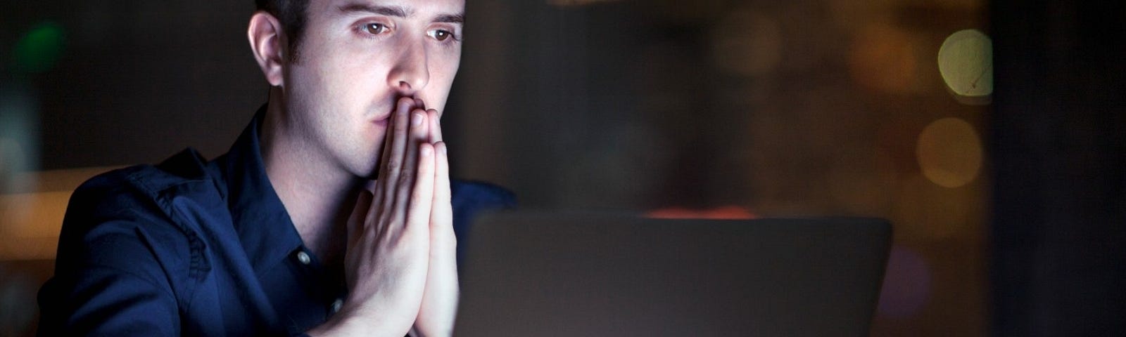 A white man sitting in a dimly lit room with a laptop on the table in front of him and his hands together in front of his mouth. Photo by Shannon Fagan/Getty Images