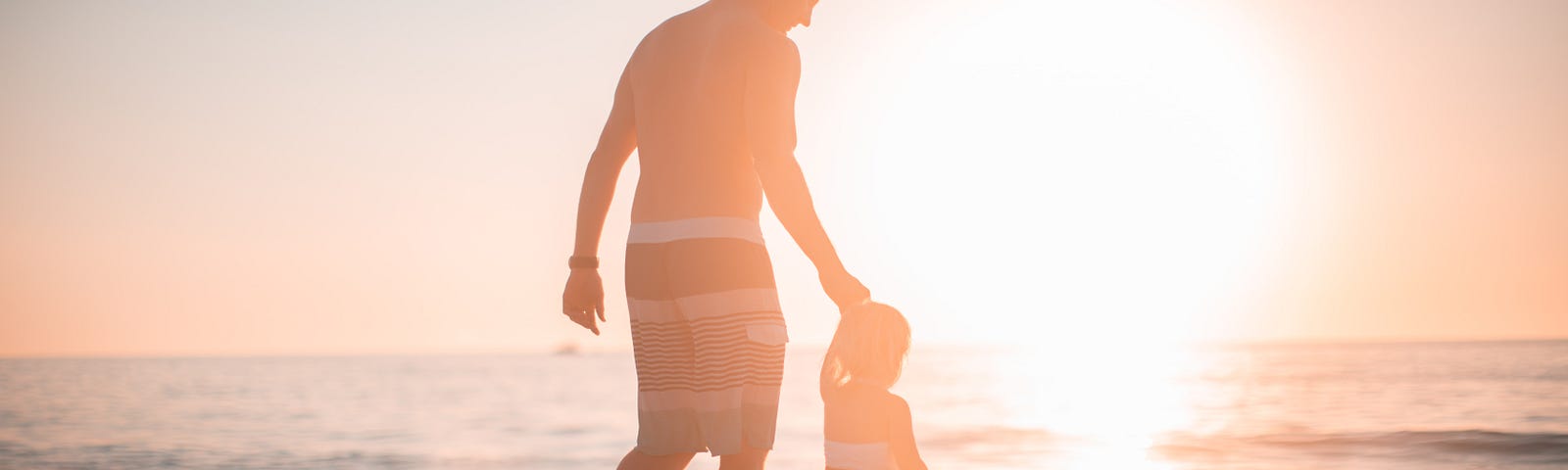 A man in swimming shorts walks hand-in-hand with a small child on a beach with a sunset in the background