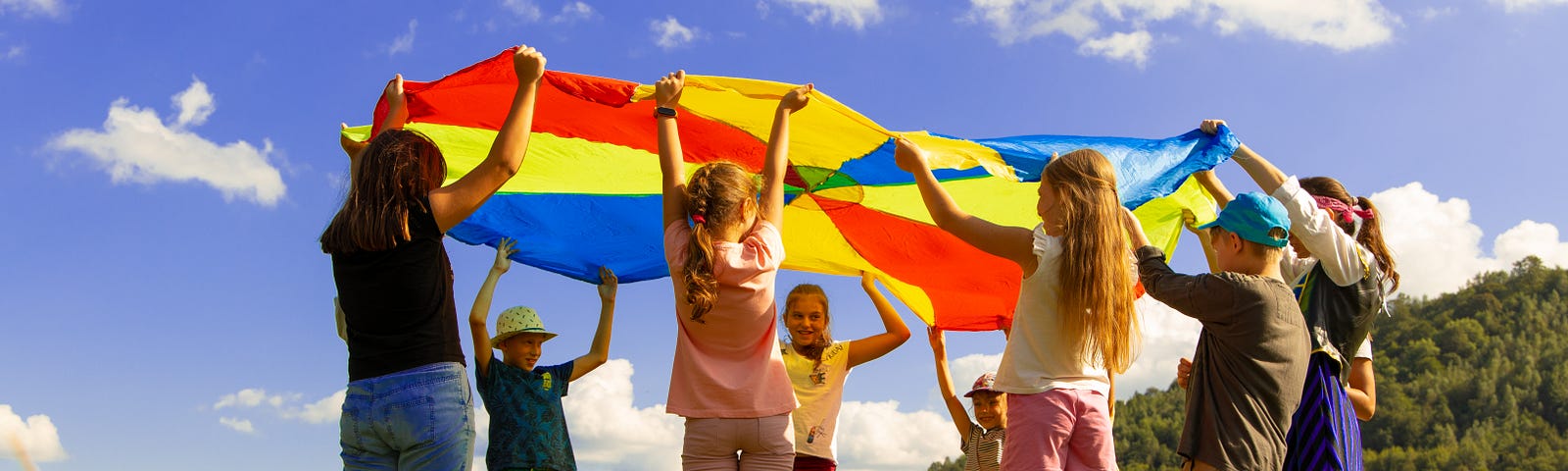 Group of people holding parachute prop up together, standing on green grass under blue skies dotted with white wispy clouds.