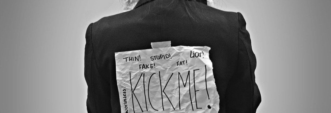 Image of a young woman wearing a Kick Me sign on her back. It symbolizes someone being bullied.