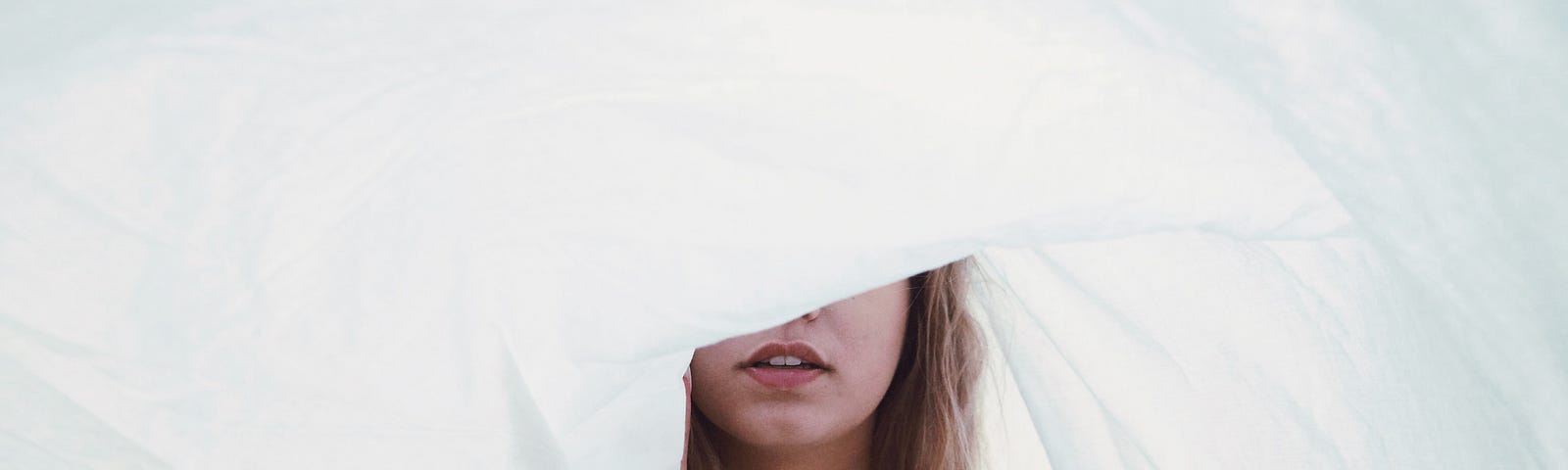 A woman, wearing a white shirt, is holding a white sheet in the air, it is covering half of her face.