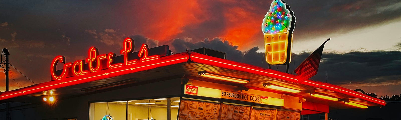 An authentic 1950’s style walk-up / drive up ice-cream parlor, with red neon lettering that says “Gabel’s” and a neon cone above more red neon piping. An American flag is barely visible as the sky darkens at sunset, with orange and dark blue clouds.