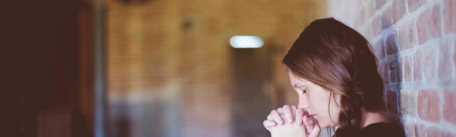 Woman leaning against a brick wall, eyes closed, her mouth resting on clasped hands