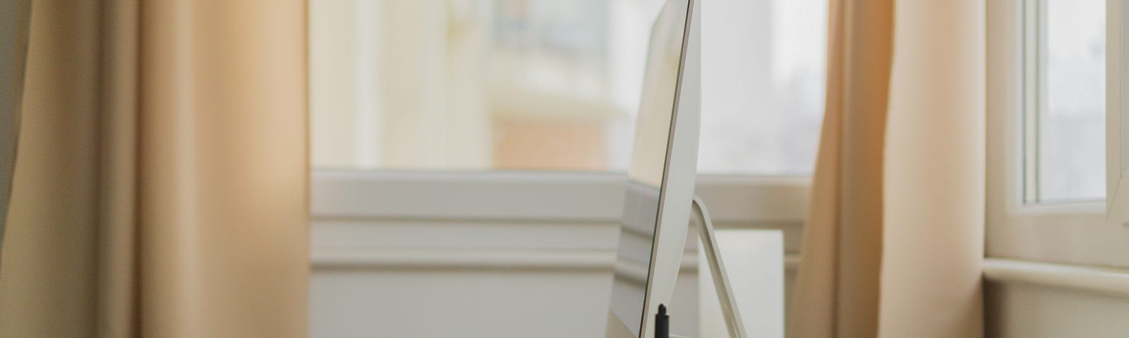 Minimalist writing setting with only a desktop computer, a keyboard, and a mouse on a beige large desk surrounded by two windows with beige curtains.