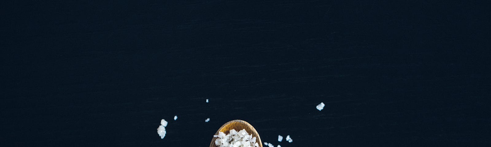 A small wooden spoon extends from the bottom of the image. The spoon has coarse salt filling it (and spilling out). There is a black background. Too much (or too little) salt is unhealthy.