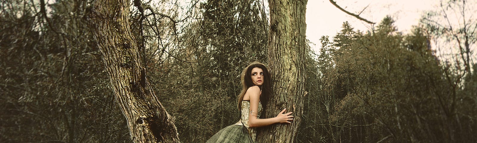 A photo of a woman hugging a tree with a long flowing green gown on with a distressed expression on her face.