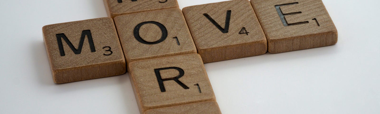 Scrabble wooden pieces, brown with black letters: The horizontal row says “MOVE” while the vertical one spells MORE. They overlap with the “O.” White background.