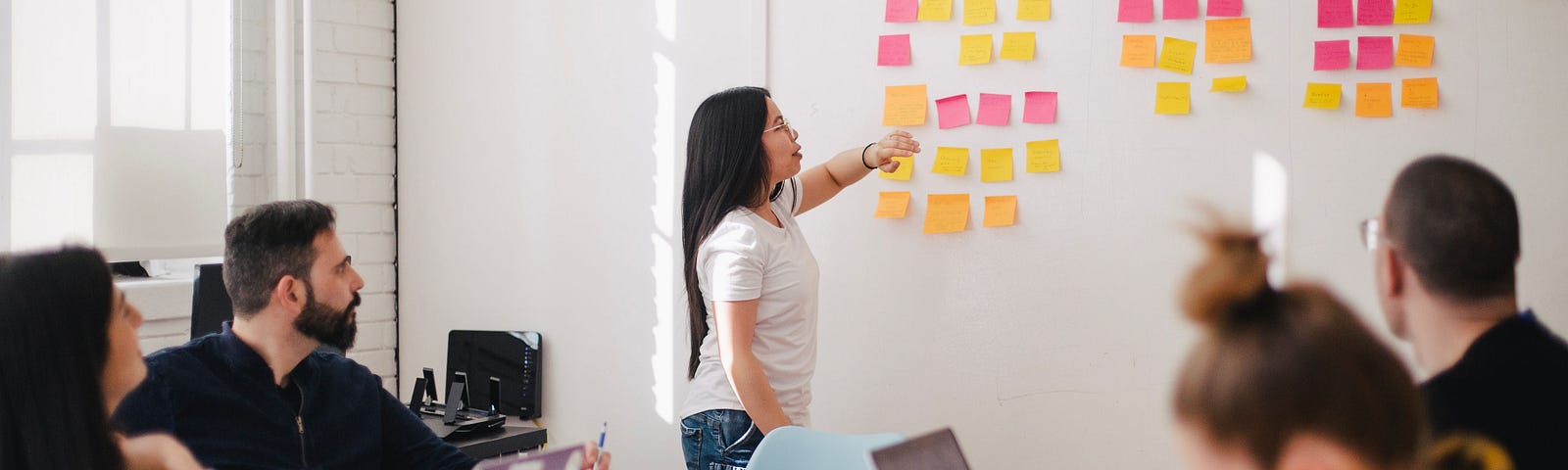 A woman stands in front of colleagues, next to a wall with sticky notes of varying colors. She points to one sticky note.