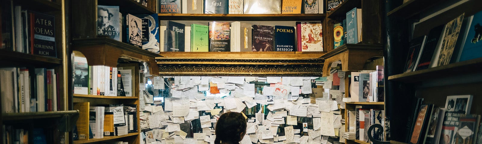 A young woman with braids stands in front of a large and very busy notice board at the back of the poetry section in a lovely old bookstore.