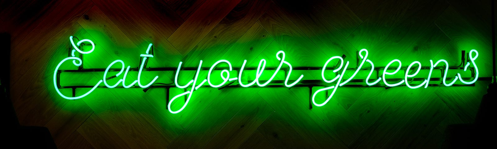 A green neon sign says Eat your greens on a dark wood chevron wall.