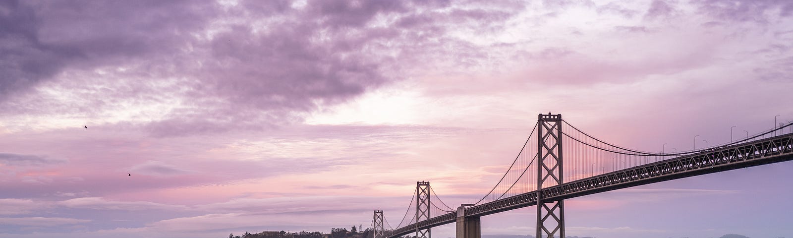 The San Francisco Bay Bridge at sunrise with a lone jogger on the shore walkway with guardrail in the foreground.