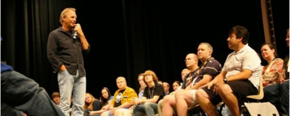 Kevin Costner speaks on a stage surrounded by students in a casual setting.