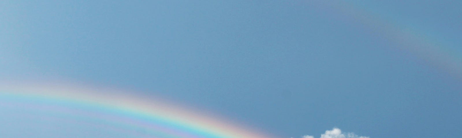A rainbow across the blue sky. There is one solitary cloud covering part of the rainbow and two treetops visible in the lower part of the photo.