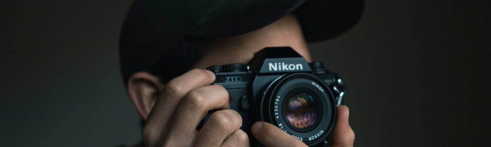 Young man wearing hat holding an old style Nikon camera up to his face to take a photo