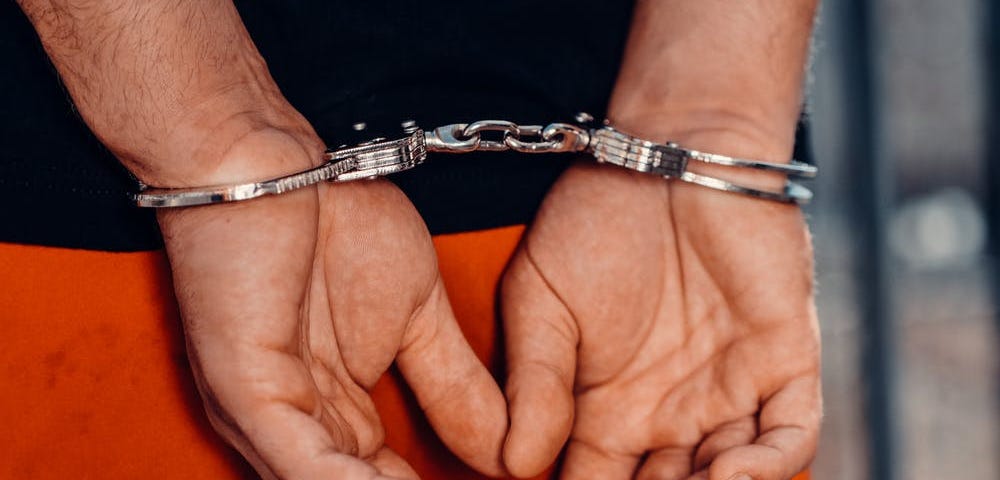 A closeup of a pair of hands in handcuffs behind a man’s back. He’s wearing a black top and orange pants.