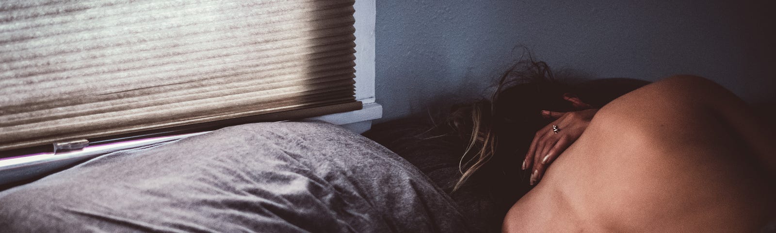 Image of a person in a bed with grey sheets, their head next to the pillow, bare back turned to the camera and a hand in their neck.