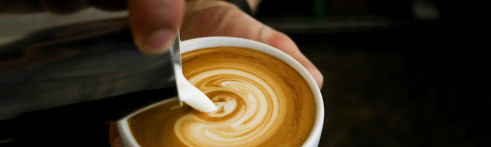 A cafe latte in someone’s left hand while the right hand pours cream.
