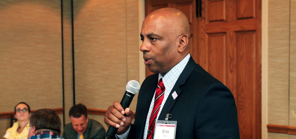 Erick Allen, dressed in a dark suit with a red tie, uses a microphone to address a room of people.