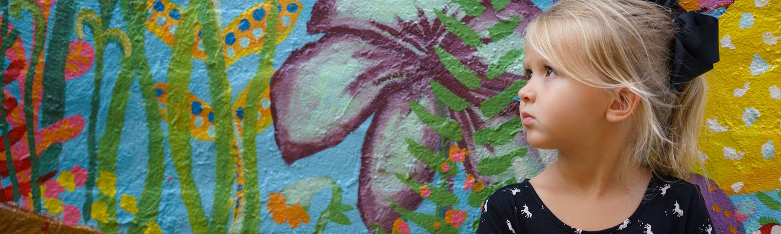 Little girl in a black dress with a small white print and her blonde hair pulled back with a large black bow, sitting in front of a colorful tropical painting.