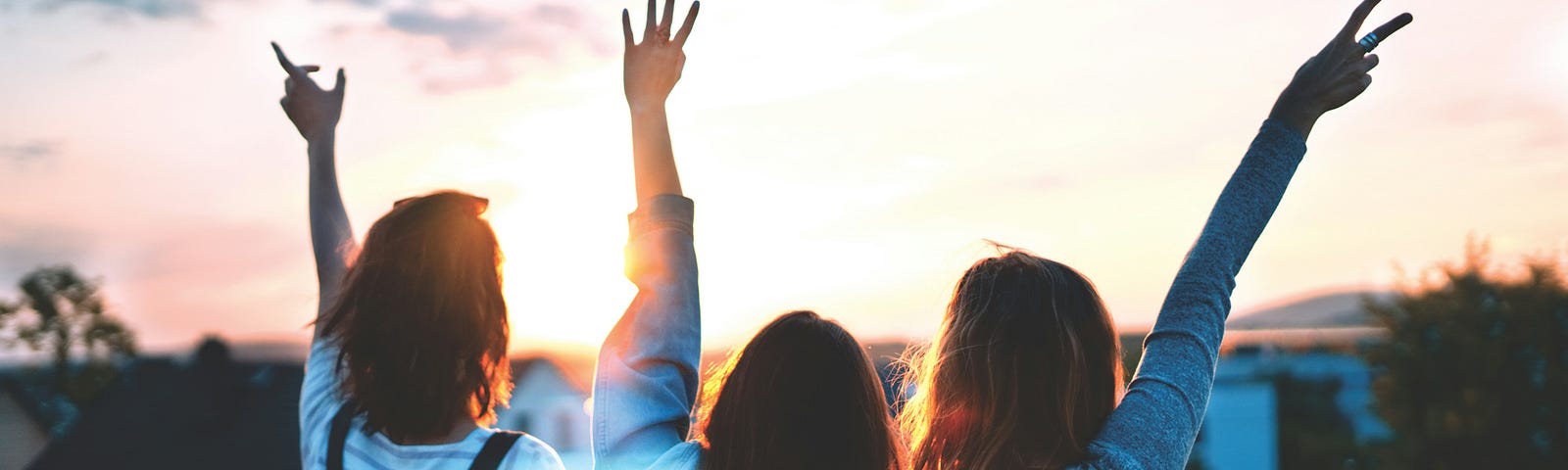 Three girls, photographed from the back raising their handsin their against a sunset.