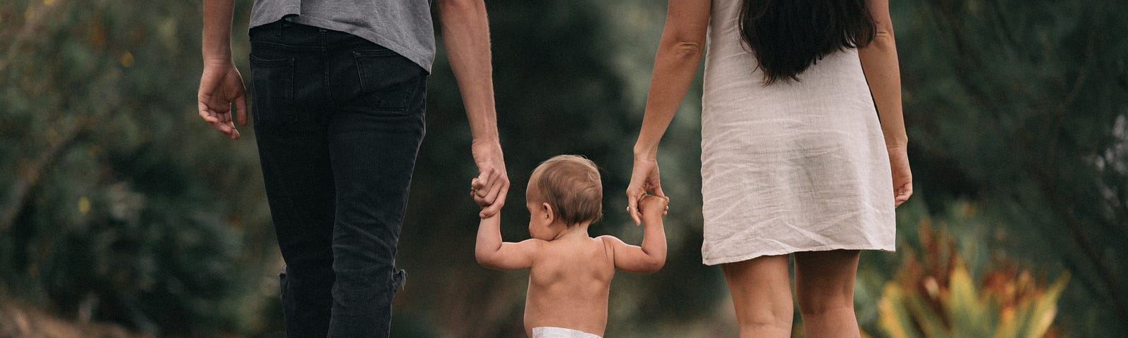 A man and a woman walking with a baby/ toddler between them, all holding hands.