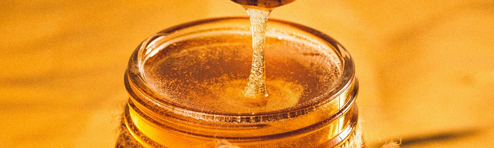 Yellow background. A big Jar full of honey. A honey dipper on top of the jar dripping honey.