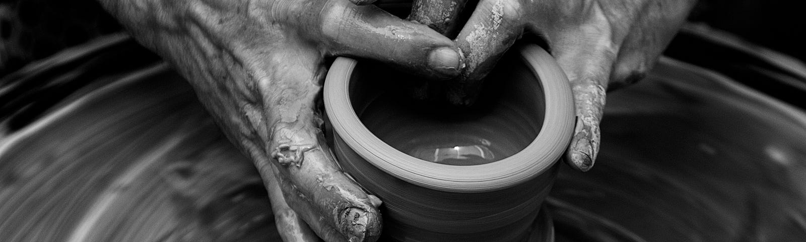 Black and white photo of two old hands shaping clay into a container while it sits on a spinning wheel.
