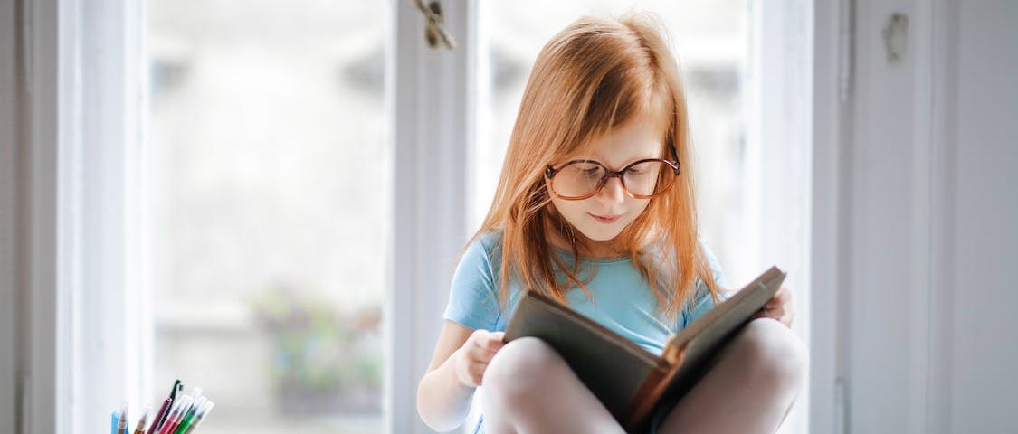 Young girl with red hear and glasses, sitting cross-legged on a window sill, reading a book.