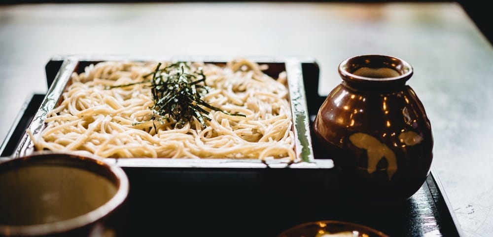 White noodles and flakes of nori in a square dish with ginger and wasabi in a smaller circular dish and two cups