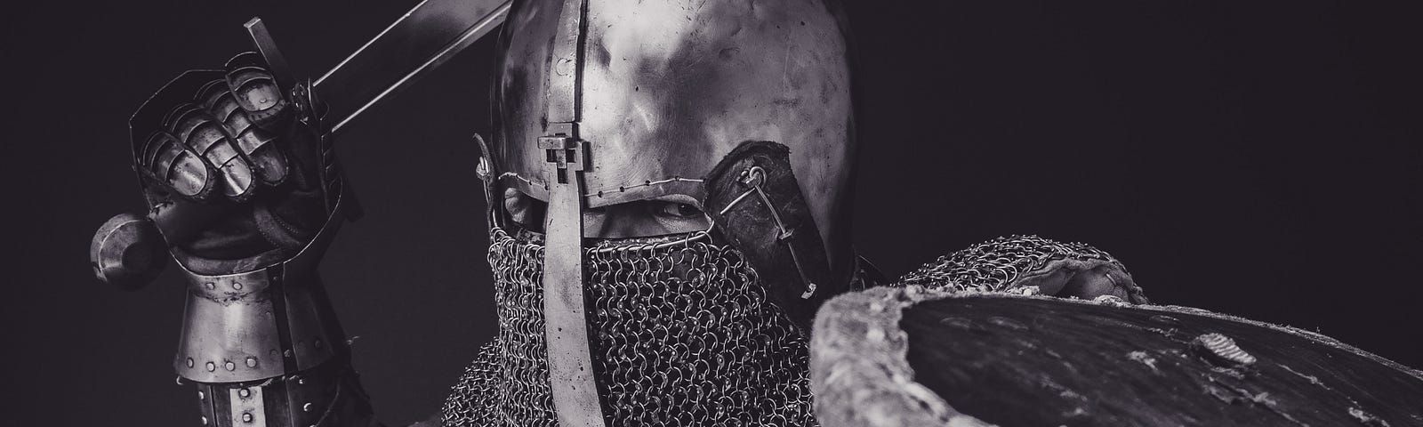 A grey-black photo of a viking-like suited solder raising sword and lifting shield aside in an attack motion right at the camera. Only the angry eyes are visible on his face which is fully covered in a metal helmet and chainmail that covers his nose and mouth.