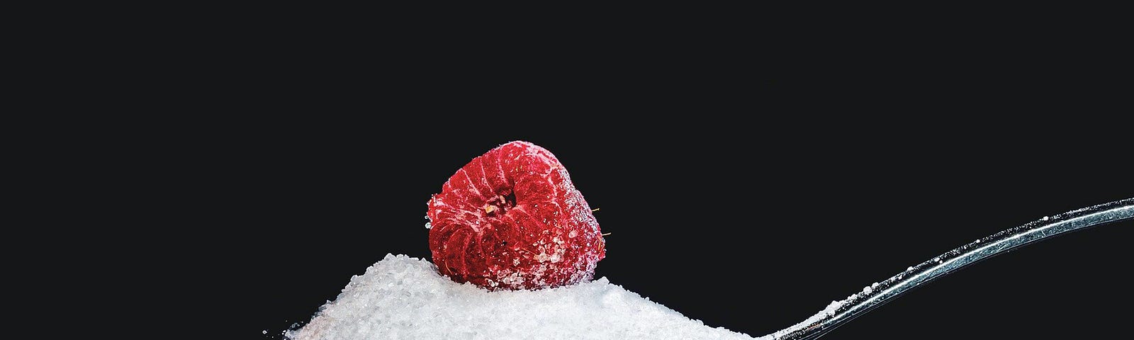 A spoon emerges from the right, heaped up with sugar (that is spilling over the sides). There is a raspberry on top of the sugar. Sugar is everywhere in America. The National Health Institute offers that adults now get 15 percent of their calories from sugar added at the table or during food processing.