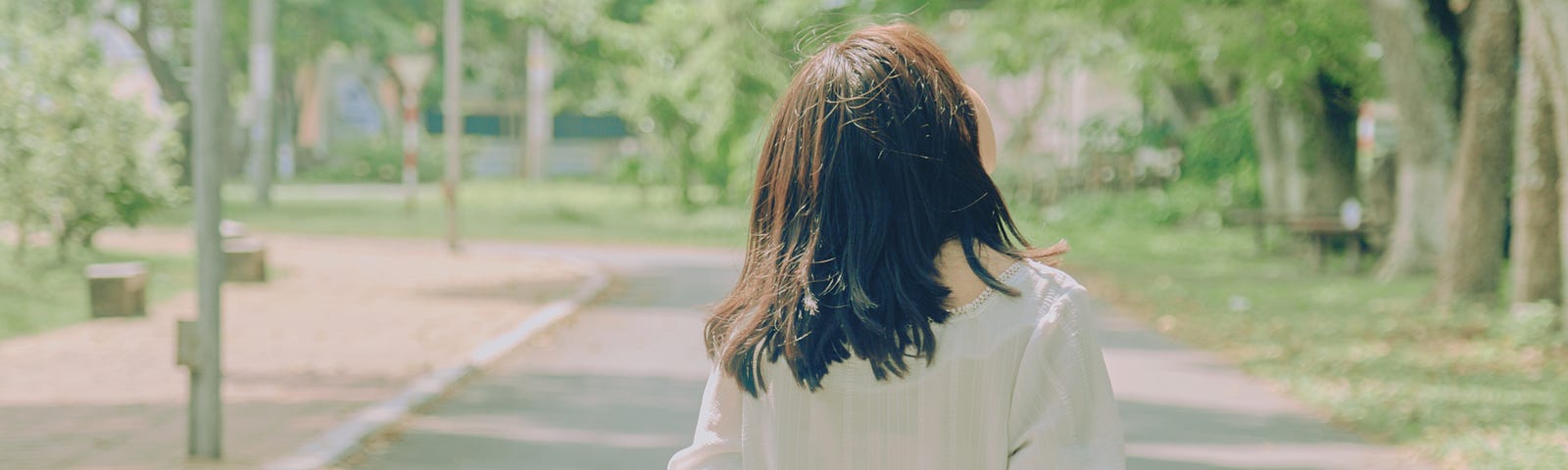 Back view of a girl holding her hands together behind her back and walking on a street among green trees.