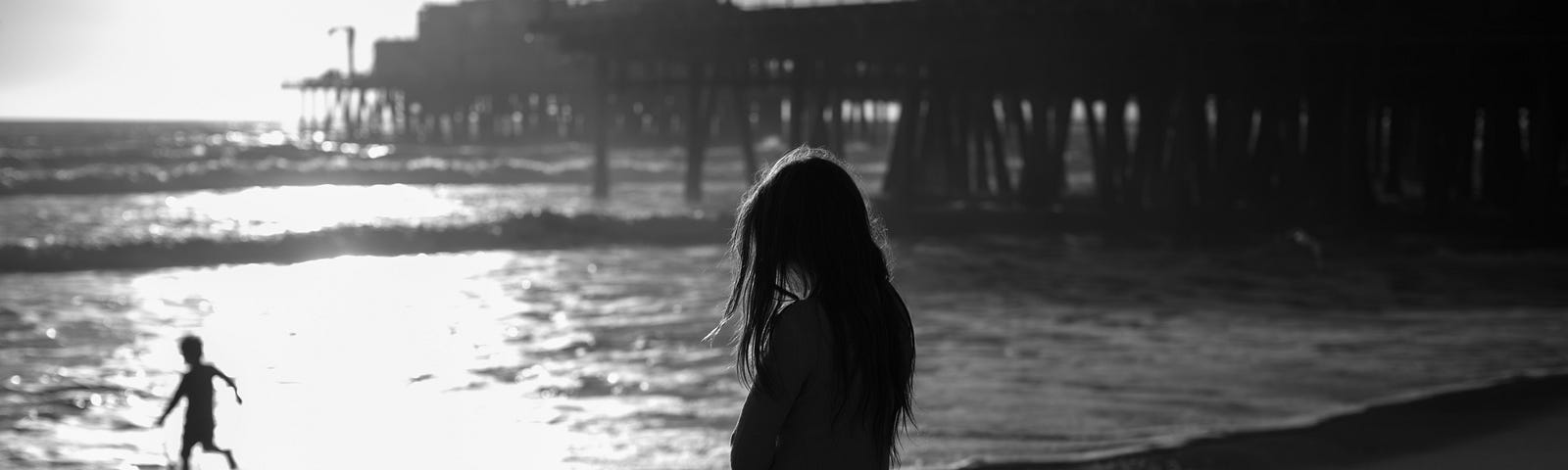 A grayscale image of a woman standing on the shore at the beach. There is also a  silhouette of a child playing in the water.