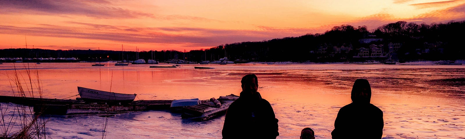 Silhouettes of three people holding hands and standing on a snowy shore during a vibrant sunset, with boats and a calm body of water in the background.