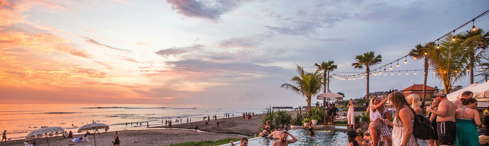 A pool full of white tourists taking pictures of themselves in front of the Balinese sunset on the beach behind them. There are palm trees and string lights framing the pool on the far right side.