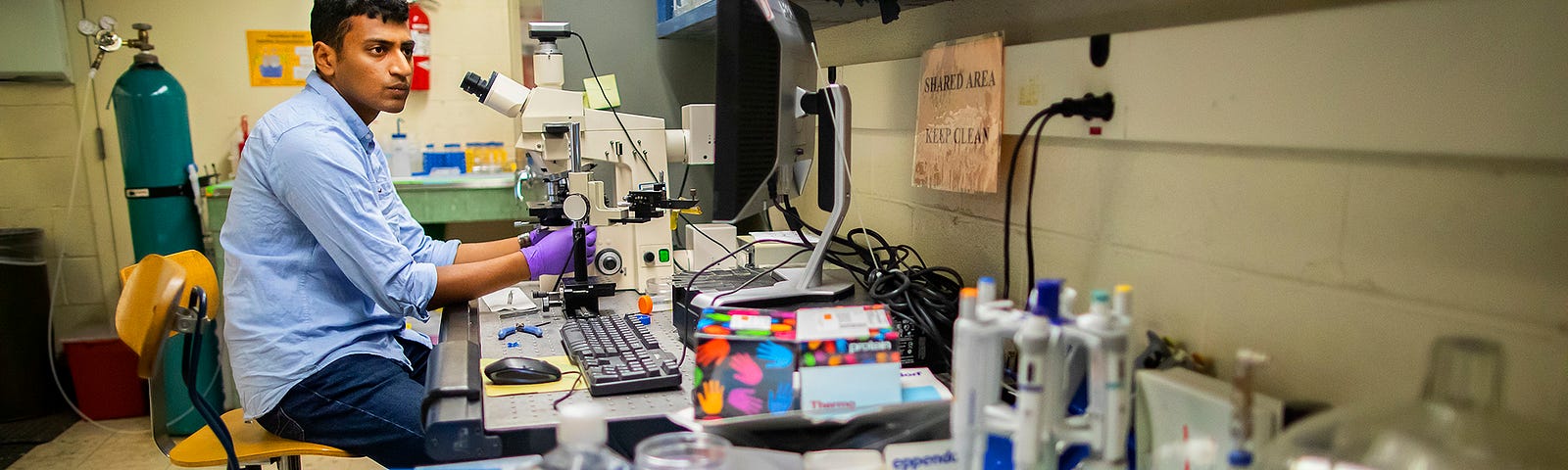 Sriram sits in front of a microscope on lab bench piled high with supplies.