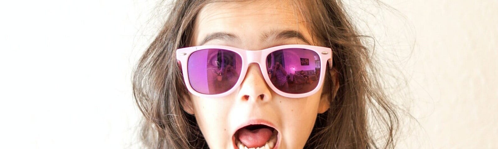 A young girl, with messy brown hair wearing reflective sunglasses, her eyebrows raised and mouth open in surprise or perhaps shock.