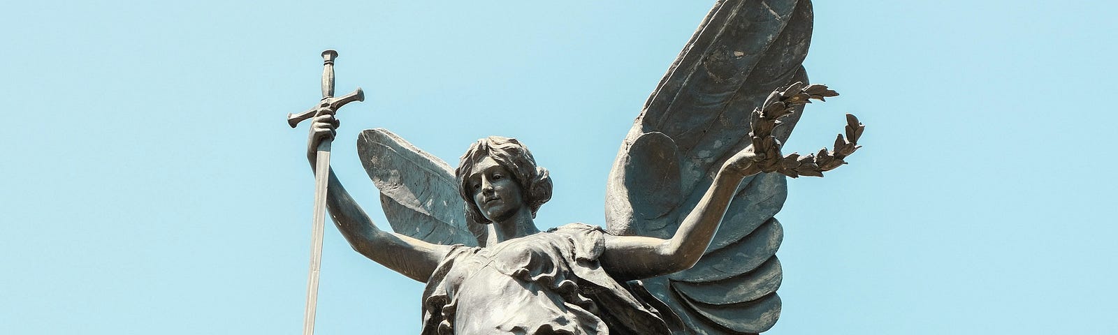 Venus, Goddess of love, a stone statue. against a hazy blue sky. She has wings and bears a sword in her right hand, and a laurel wreath in her lest